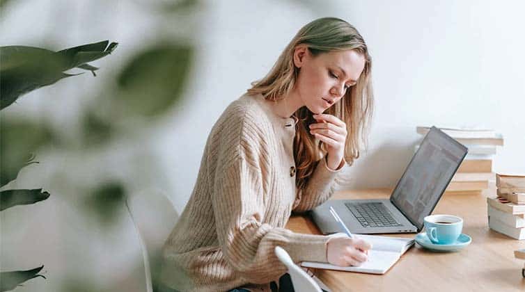 Woman taking notes at table with laptop and cup of coffee