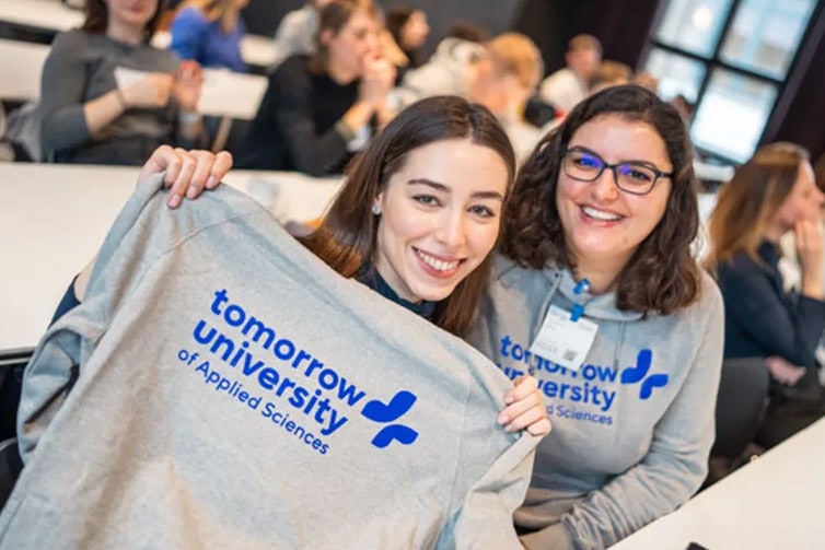 Image of two students sat at a table holding up a grey jumper that says 'Tomorrow University of Applied Sciences' on it