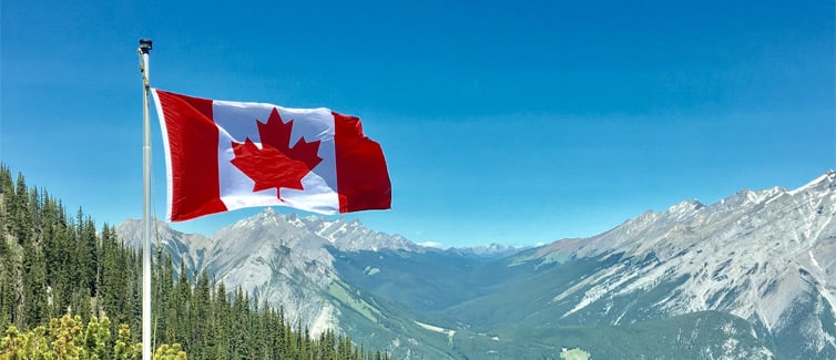 Image of the Canadian flag on a flagpole flying in front of a mountain range and a forest of trees