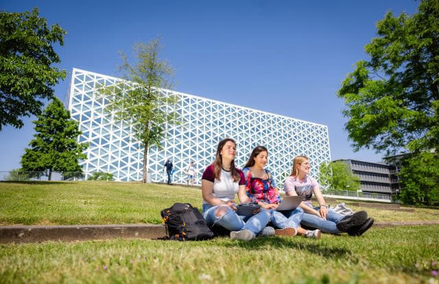 Image of three students sat down on grass in front of a big glass building