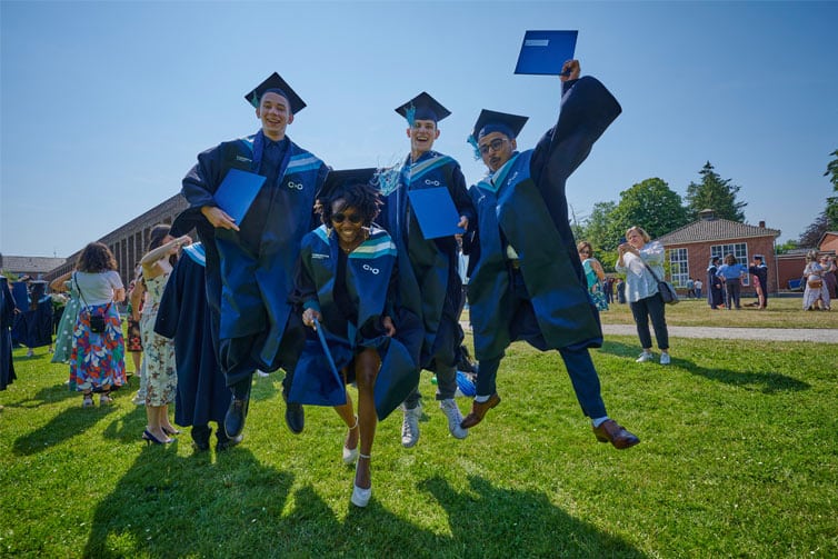 4 students wearing blue graduation gowns and caps, jumping up in the air, smiling at the camera