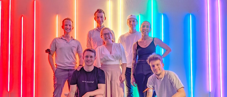 Image of 7 students standing in front of rainbow neon lights smiling at the camera