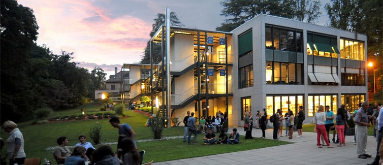 Students standing and sitting around tables outside of a big white and glass building