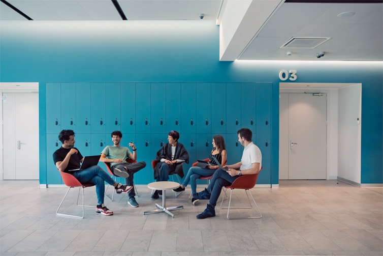 Image of students sitting on chairs in a hallway in front of a blue wall