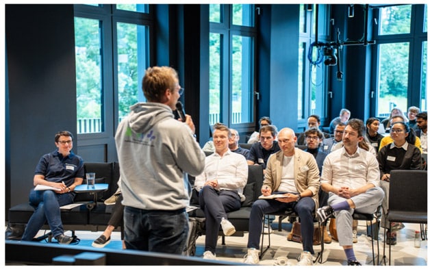 Students sat on chairs facing towards a person using a microphone to speak with their back to the camera