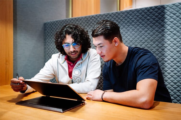 Two students sat at a table looking at a laptop screen together