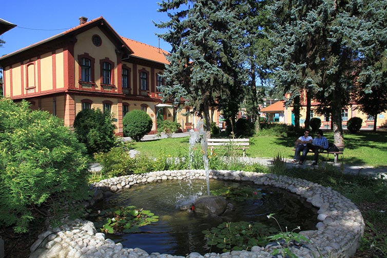 Image of a red and yellow building behind a pond with a water fountain in it, surrounded by trees